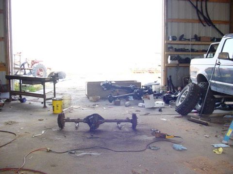 1992 Ford Bronco - A 1992 Ford Bronco is partially visible in a garage, surrounded by vehicle axles and tools.