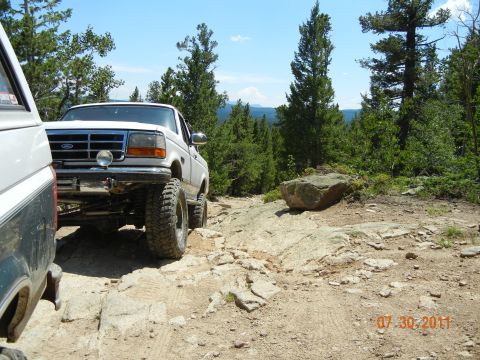1996 Ford Bronco - Two trucks navigate a challenging, rocky mountain trail under a clear blue sky.
