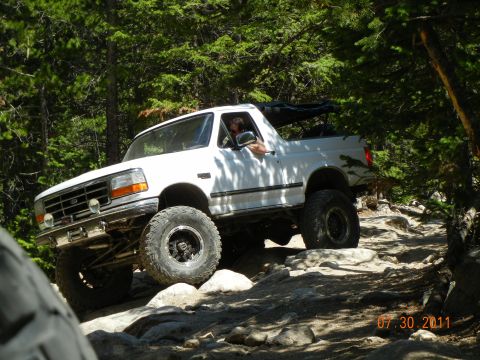 1996 Ford Bronco - A white 1996 Ford Bronco tackles a challenging rocky path in a wooded area.