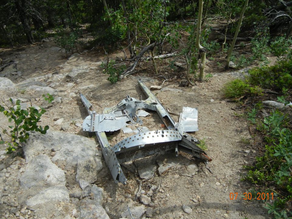 Airplane wreckage on rocky, wooded terrain. Metal debris is scattered and broken