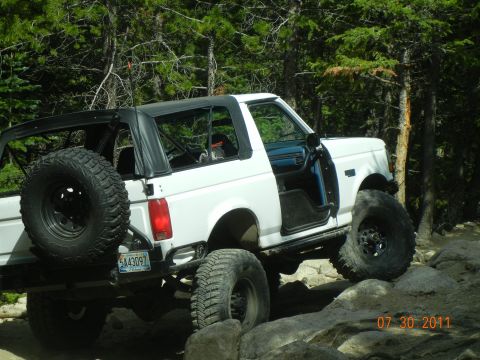 1996 Ford Bronco - White Ford Bronco with soft top and spare tire on rear, driving over rocks on a trail.