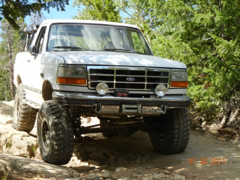 1996 Ford Bronco - A white Ford Bronco navigates a rough, wooded terrain with its front end prominently displayed.