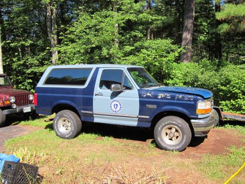 1991 Ford Crown Victoria LTD - This two-tone blue Ford Bronco is parked outdoors next to a red Jeep, with trees in the background.