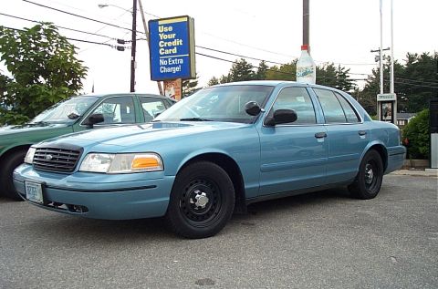 2001 Ford Crown Victoria - A blue 2001 Ford Crown Victoria sedan is parked next to another car, with a sign in the background.