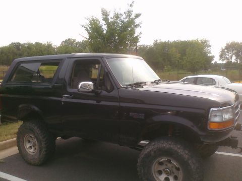 1995 Ford Bronco - This lifted black Ford Bronco is parked in a lot, with trees and a fence in the background.