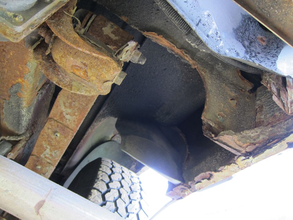 Undercarriage of a heavily rusted vehicle, showing leaf springs, a tire, and extensive corrosion on frame components