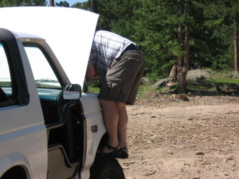 1995 Ford Bronco - A person in a plaid shirt and cargo shorts leans deep under the raised hood of a white full-size Ford truck parked on a dirt clearing in a…
