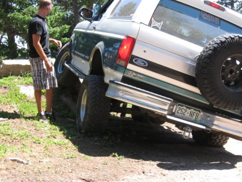 1995 Ford Bronco - Low three-quarter rear view of a green and silver two-tone Ford Bronco flexing over a rock ledge on a wooded trail, one rear wheel lifted a…