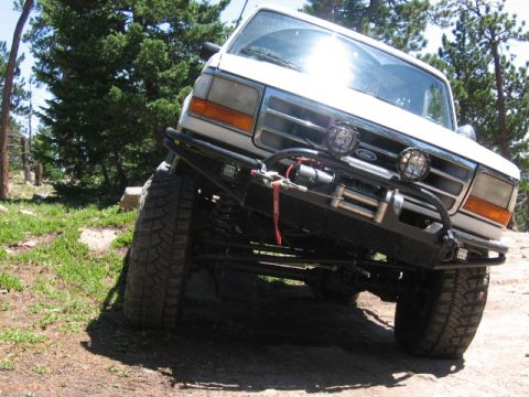 1995 Ford Bronco - Low front view of a white Ford 4x4 with a black tube bumper carrying a mounted winch and round auxiliary lights, riding on mud-terrain tire…