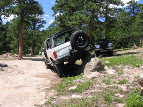 1995 Ford Bronco - A silver full-size Ford Bronco with its hardtop removed and a spare tire mounted on a swing carrier at the rear, climbing over a boulder on…