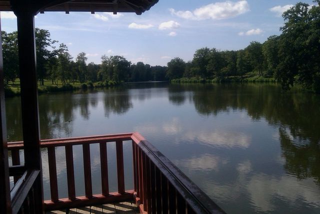 Wooden railing and gazebo overlooking a calm lake with trees on the far bank under a cloudy sky