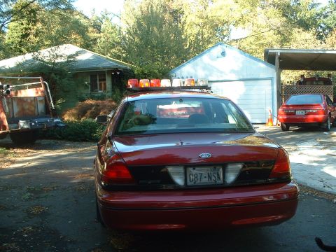 1999 Ford Crown Victoria - A red 1999 Ford Crown Victoria, possibly a police car restoration project, is parked in a driveway with another car visible.