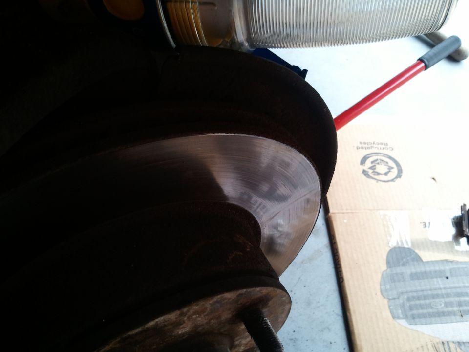 Car brake rotor with visible wear marks, caliper partially visible, headlight assembly above