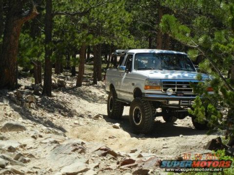 1995 Ford Bronco - A white 1990s Ford Bronco full-size SUV with a lift kit and mud-terrain tires crawling over a rocky trail on a wooded off-road path. The tr…