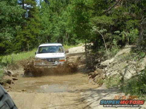 1995 Ford Bronco - A white Ford Bronco splashes through a muddy water crossing on a wooded dirt trail, kicking up brown water and mud from its front tires. Th…