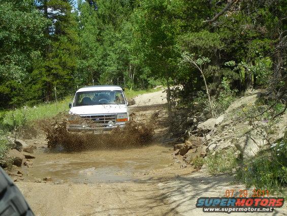 White Ford Bronco splashing through mud puddle on wooded off-road trail