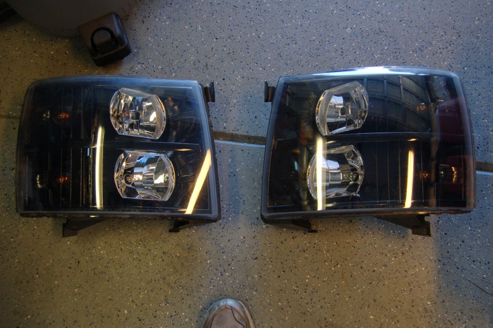 Two black aftermarket headlights, likely for a truck, sitting on a speckled floor