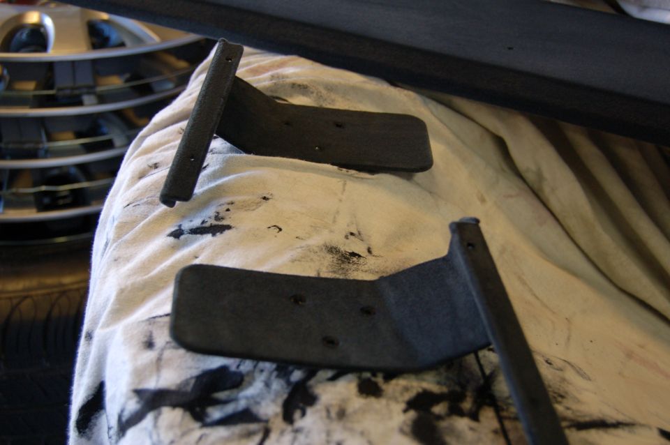 Two black metal shelf brackets with screw holes on a white cloth