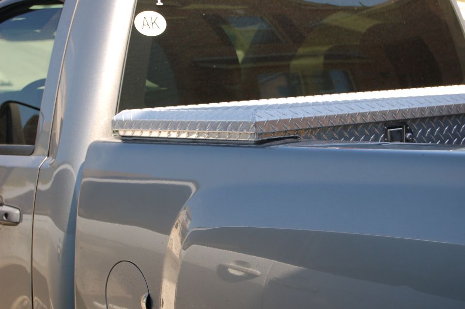 Side view of a truck bed with a diamond plate toolbox on top. There's an 'AK' sticker on the rear window