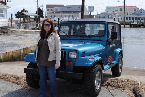 1991 Jeep YJ - A woman stands next to a blue 1991 Jeep YJ in a coastal town with houses in the background.