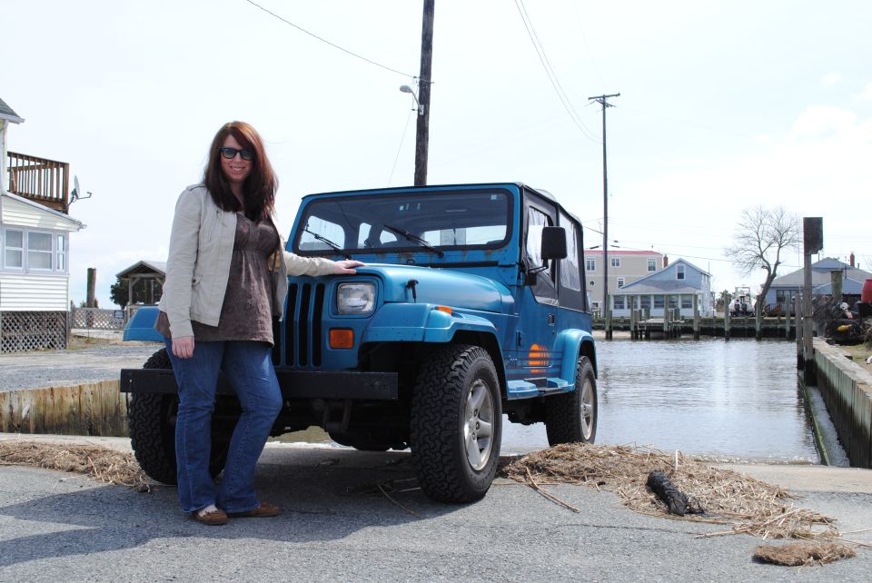 Blue 1991 Jeep YJ parked on a boat ramp next to water, with a woman standing beside it