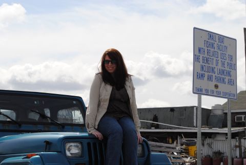 1991 Jeep YJ - A woman poses on the hood of a blue Jeep Wrangler with a sign for a small boat fishing facility behind her.
