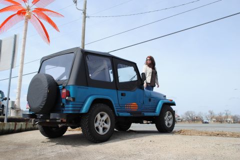 1991 Jeep YJ - A woman stands by a blue 1991 Jeep YJ with a black soft top, parked outdoors near a body of water.
