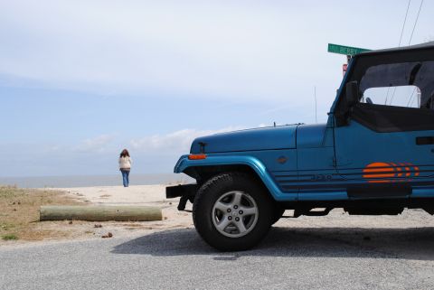 1991 Jeep YJ - A blue Jeep Wrangler is parked on the side of the road next to a beach with a person walking in the distance.