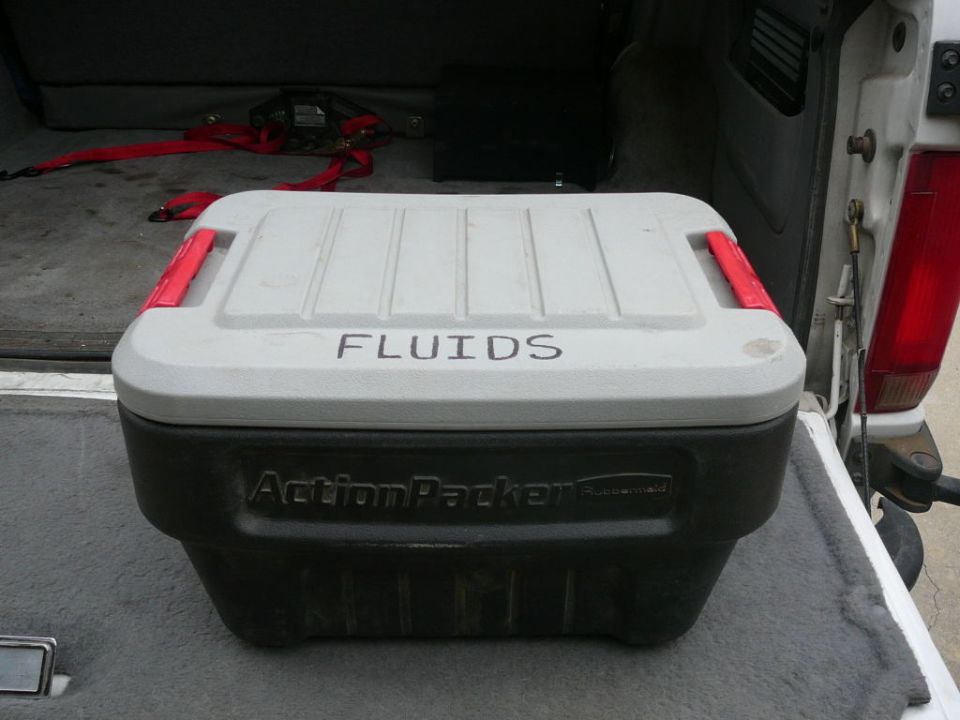 Gray and black ActionPacker storage container with 'FLUIDS' label in a vehicle cargo area