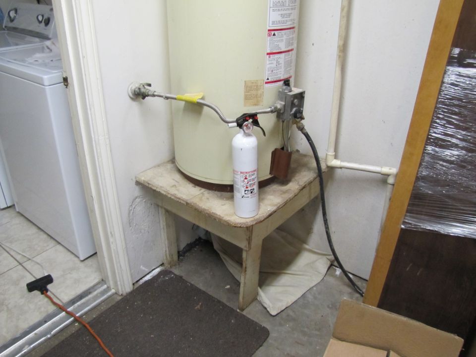 Water heater on a wooden stand with a fire extinguisher and plumbing. A washing machine is visible to the left