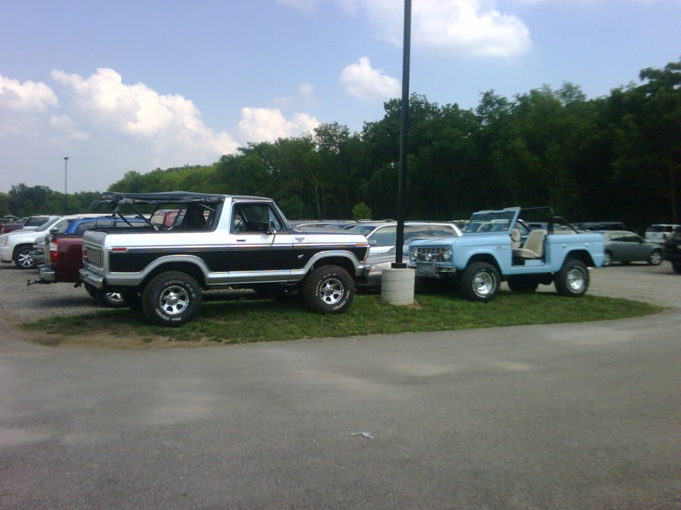 Two SUVs, a white and black Bronco and a light blue Bronco, parked on grass. The white Bronco is a 1978 model