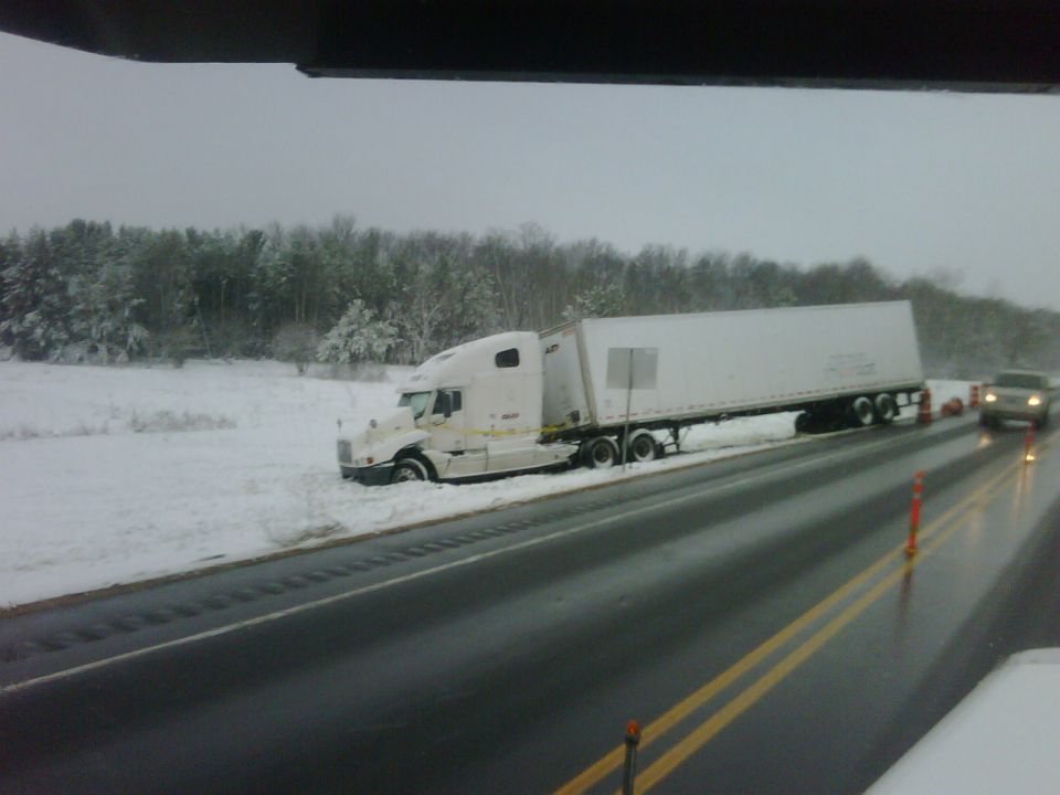Semi-truck with trailer stuck in snow beside a highway, with another vehicle visible on the wet road