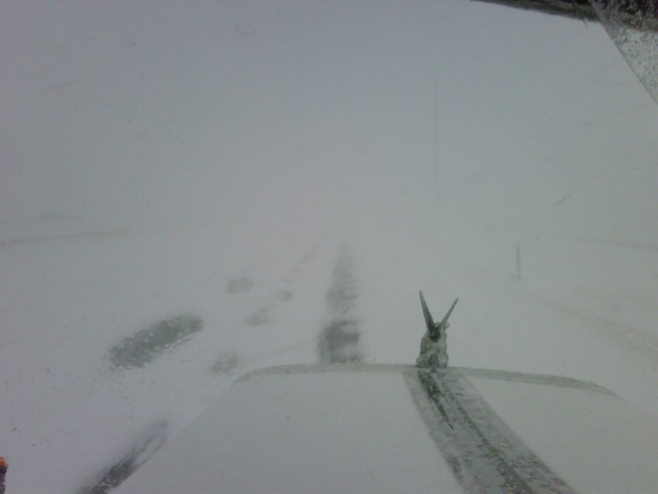 View from inside a vehicle driving on a snowy road with tracks visible ahead. A small object with two points sticks up from the dashboard