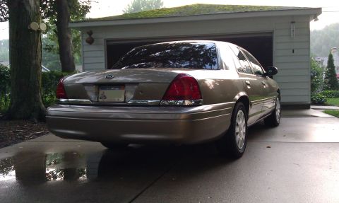 2007 Ford Crown Victoria - The rear of a gold sedan is parked on a wet driveway in front of an open garage door.