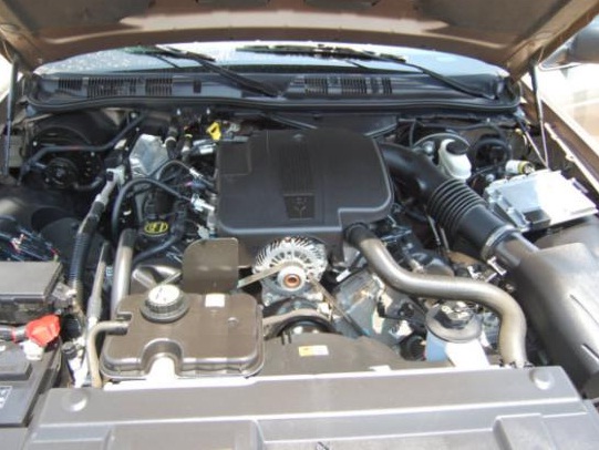Open engine bay showing black engine cover, belt pulleys, intake tubing and battery.