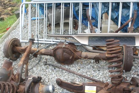 1996 Ford Bronco - A heavily rusted rear axle assembly, complete with springs and brake drums, lies on a gravel surface. The Supermotors.net logo is visible i…