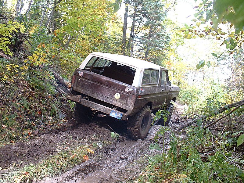 1978 Ford Bronco navigating a muddy, wooded trail with trees and fallen logs