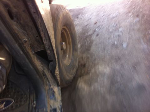 1990 Ford Bronco - A low-angle view of a vehicle's undercarriage and tire on a dusty, off-road path. The SuperMotors.net logo is visible in the lower right co…