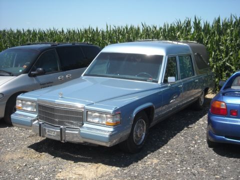 1991 Ford LTD Crown Victoria - A light blue hearse is parked outdoors next to other vehicles, with a cornfield in the background.