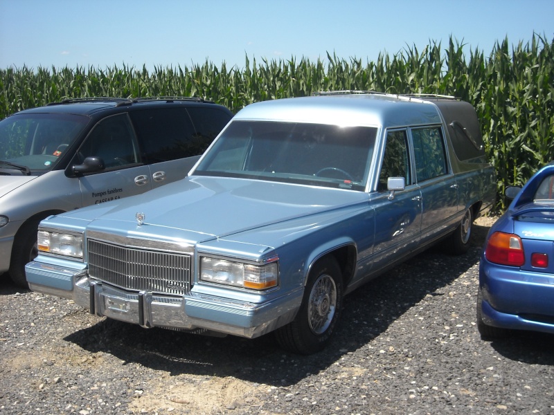Light blue hearse with chrome grille and trim, parked next to a silver minivan and a blue sports car