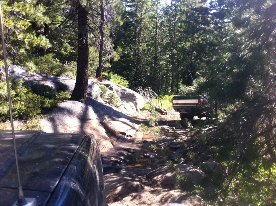 Two off-road vehicles navigating a rough, wooded trail with rocks and puddles