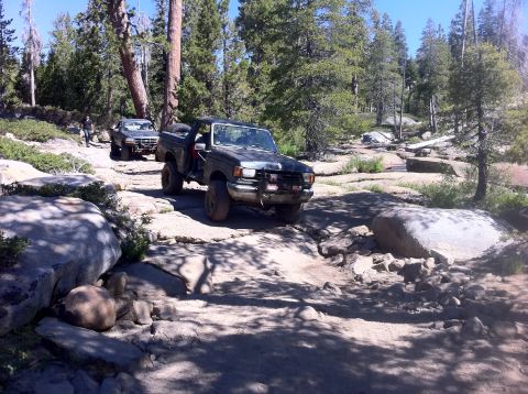 1990 Ford Bronco - Two Ford F-Series trucks traverse a challenging rocky path in a wooded area.