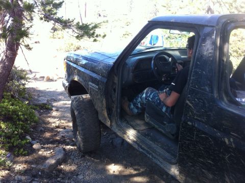 1990 Ford Bronco - A person sits in the driver's seat of a dirty Ford Bronco on a rocky, wooded trail.
