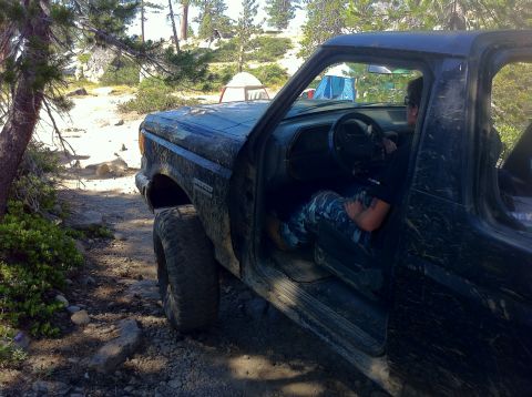 1990 Ford Bronco - A muddy Ford Bronco is parked on a dirt trail, with tents visible in the distance.