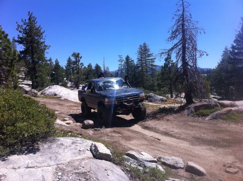 1990 Ford Bronco - A modified Ford F-Series truck navigates a rocky off-road trail under a clear blue sky.
