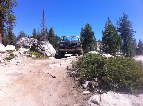 1990 Ford Bronco - A dark Ford Bronco navigates a challenging rocky trail under a clear blue sky.