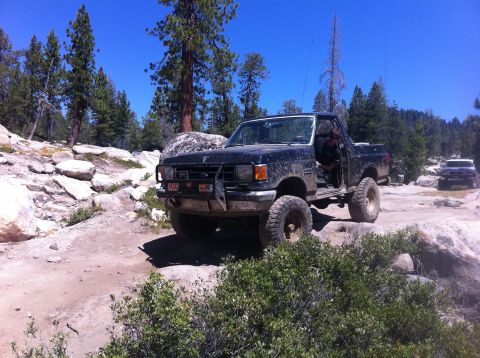 1990 Ford Bronco - A black Ford F-Series truck navigates a rocky, off-road trail under a clear blue sky.