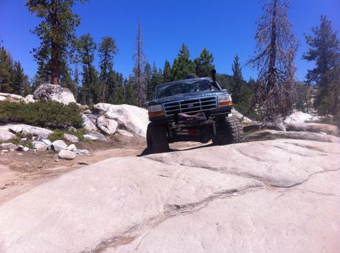 1990 Ford Bronco - A lifted Ford Bronco navigates a rocky off-road trail under a clear blue sky.