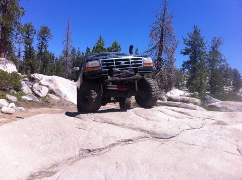 1990 Ford Bronco - A modified Ford Bronco navigates a challenging rocky terrain under a clear blue sky.