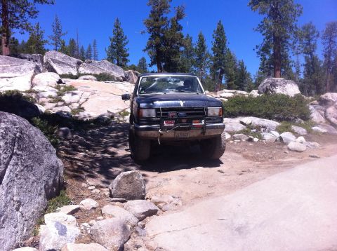1990 Ford Bronco - A dark blue Ford Bronco navigates a rocky, off-road path under a clear blue sky.